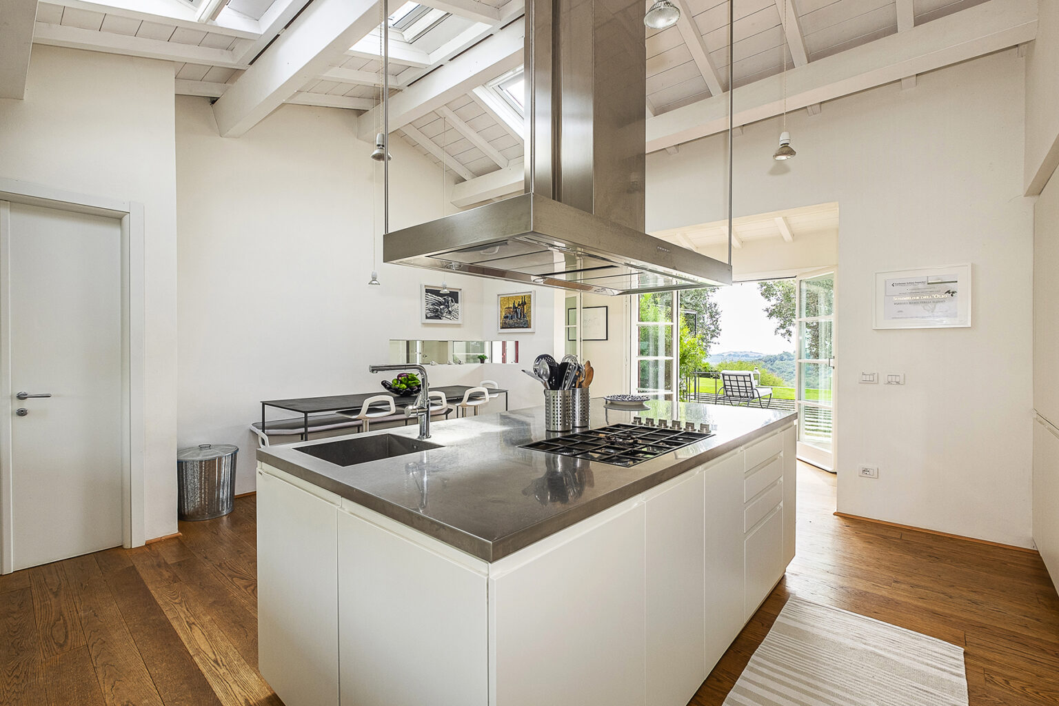 Modern kitchen with high ceilings, stainless steel appliances, and a spacious island. Features include a gas cooktop, sleek cabinetry, and large windows providing natural light and scenic views. The adjacent dining area offers minimalist furniture and a cozy atmosphere. | Only on SopranoVillas
