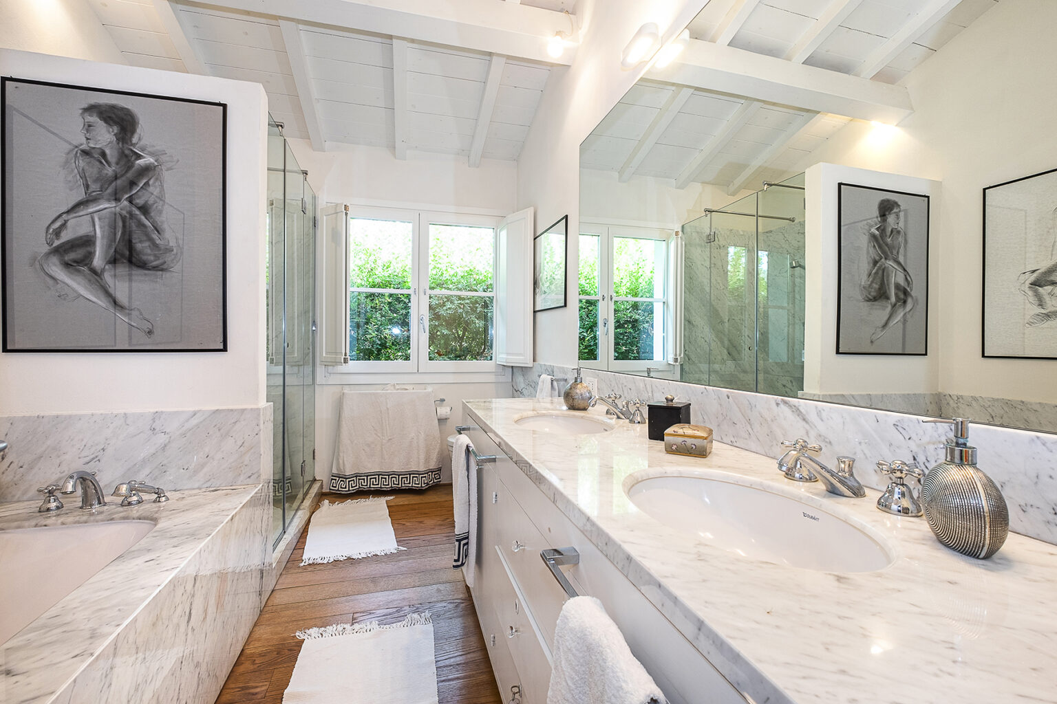 Modern bathroom featuring a marble countertop, dual sinks, and elegant fixtures. Natural light streams through large windows, highlighting contemporary artwork on the walls. The space includes a glass shower and wooden flooring, creating a stylish and serene atmosphere. | Only on SopranoVillas