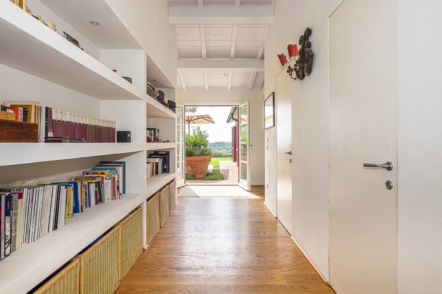 Bright and airy hallway featuring white walls, wooden flooring, and built-in bookshelves filled with books and decorative items. The doorway at the end opens to a scenic outdoor area with a potted plant and patio umbrella, showcasing a blend of indoor and outdoor living. | Only on SopranoVillas
