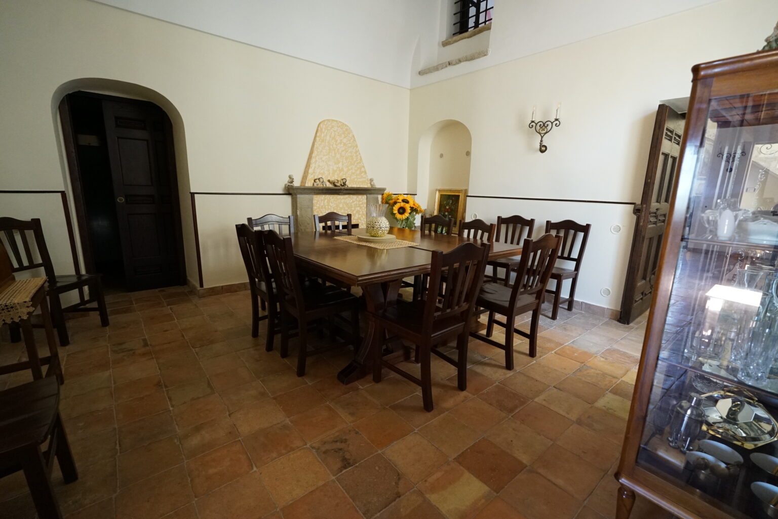 Spacious dining room featuring a wooden table surrounded by matching chairs, with a rustic ambiance created by terracotta flooring and soft wall colors, complemented by a glass display cabinet and natural light. | Only on SopranoVillas