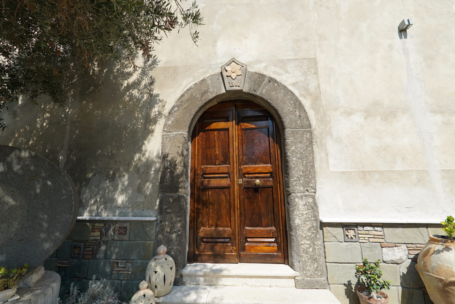 Wooden door with stone archway and decorative elements, set against a textured wall, surrounded by greenery and ornamental pots. Ideal for showcasing architectural details and outdoor design. | Only on SopranoVillas
