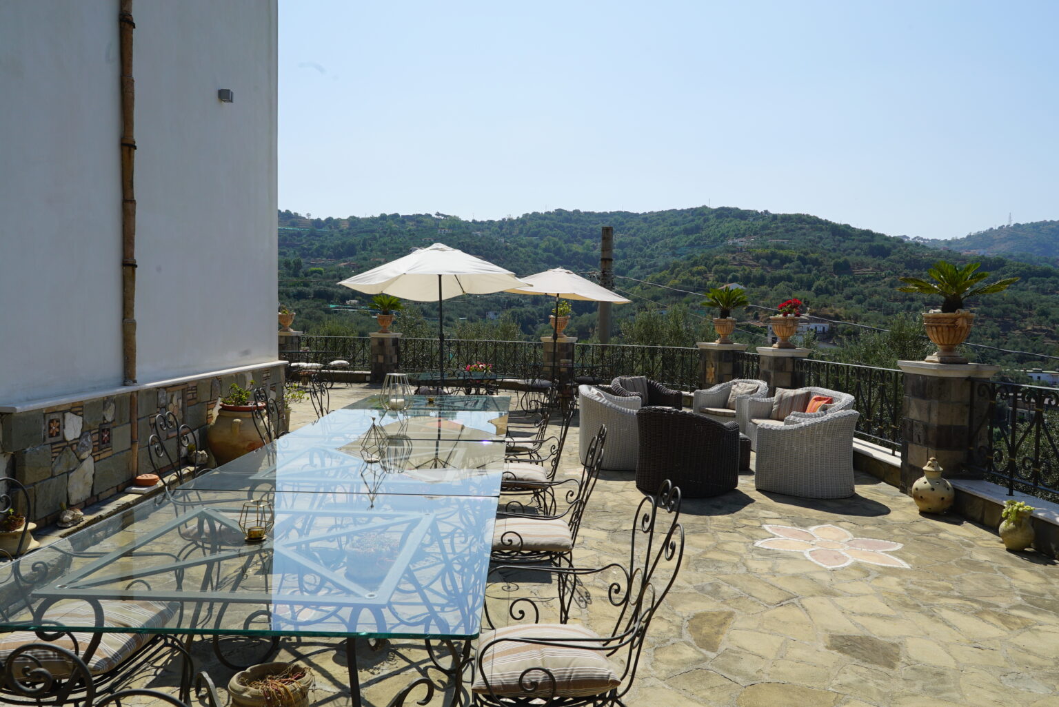 Spacious outdoor terrace with a glass dining table, comfortable seating, and umbrellas, offering scenic views of rolling hills and greenery. Ideal for relaxation and outdoor dining. | Only on SopranoVillas