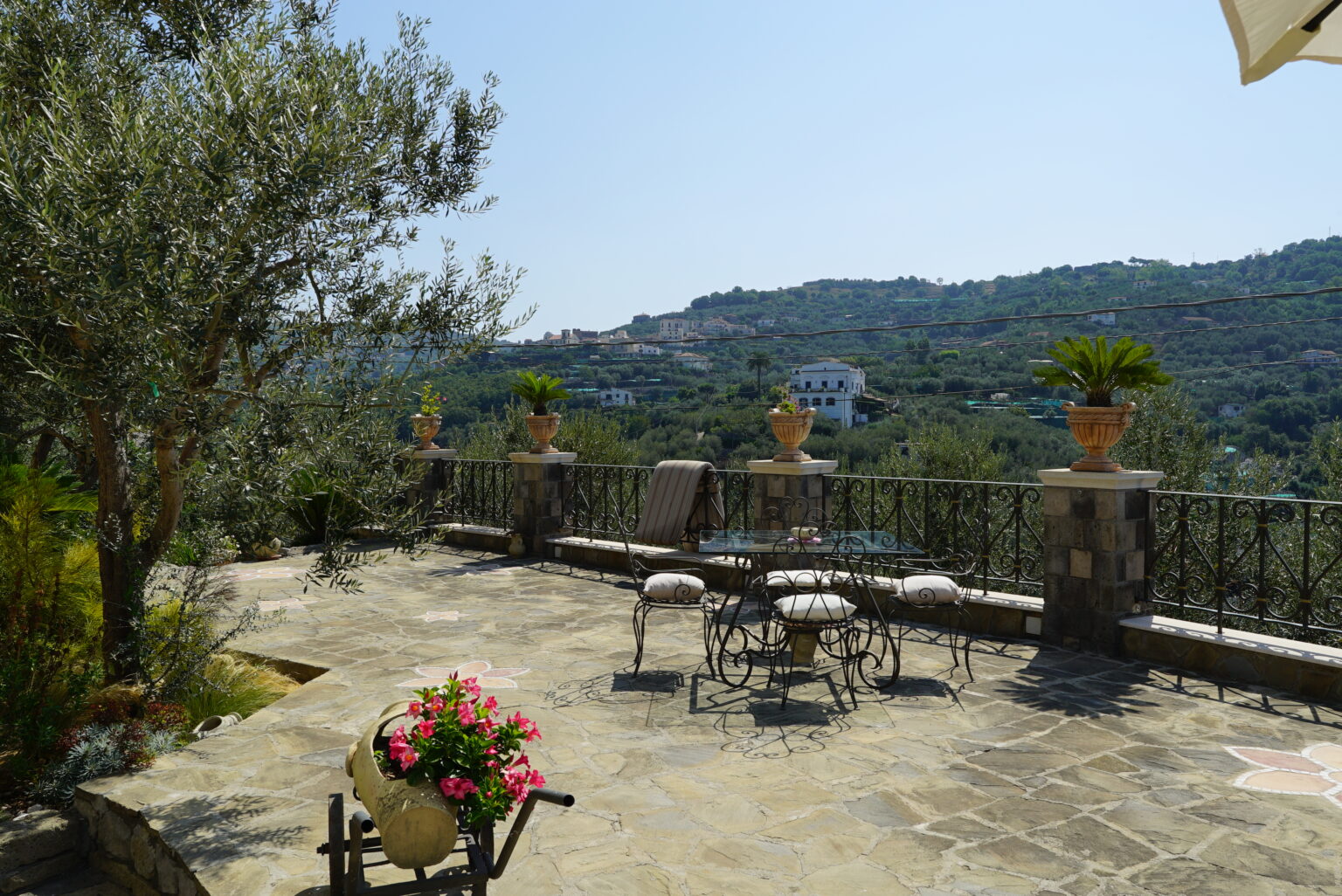 Scenic terrace with stone flooring, surrounded by lush greenery and potted plants, offering a panoramic view of hills and distant houses under a clear blue sky. | Only on SopranoVillas