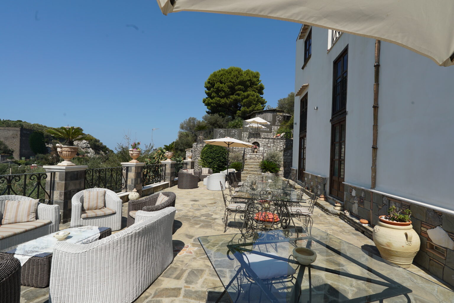 Outdoor patio area with stylish wicker furniture, a glass table, and potted plants, set against a clear blue sky and scenic landscape. Perfect for relaxation and entertaining. | Only on SopranoVillas