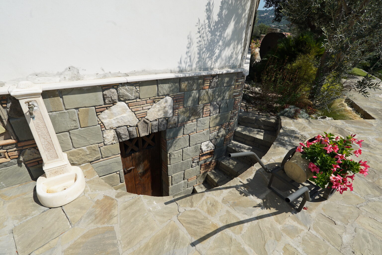 Stone staircase leading to a rustic wooden door at the base of a house, surrounded by natural stone walls and a vibrant flower pot, showcasing a serene outdoor setting. | Only on SopranoVillas