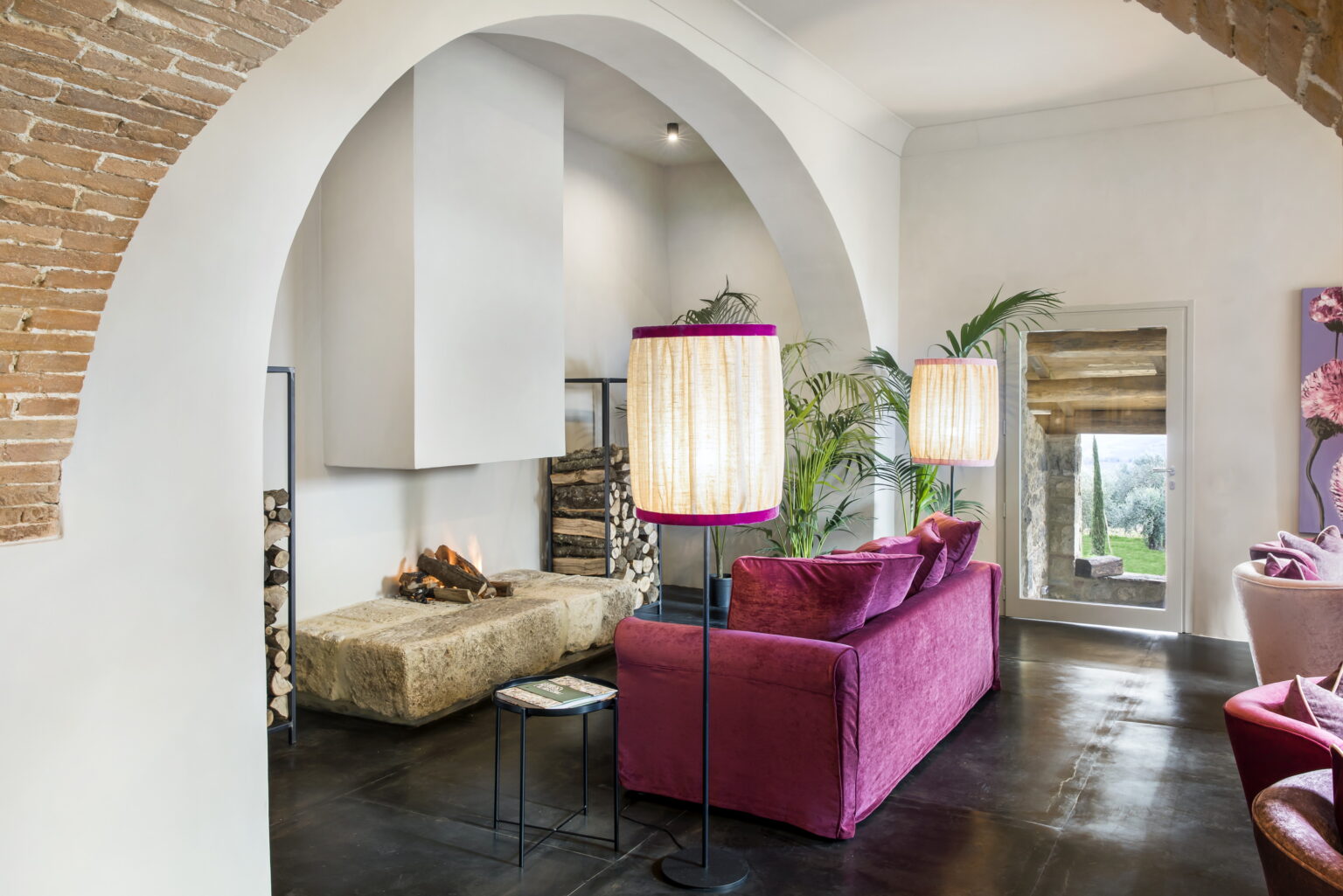 Modern living room with arched architecture featuring vibrant pink sofas, stylish floor lamps, and a cozy fireplace. Large windows provide natural light and a view of greenery, enhancing the contemporary decor. | Only on SopranoVillas