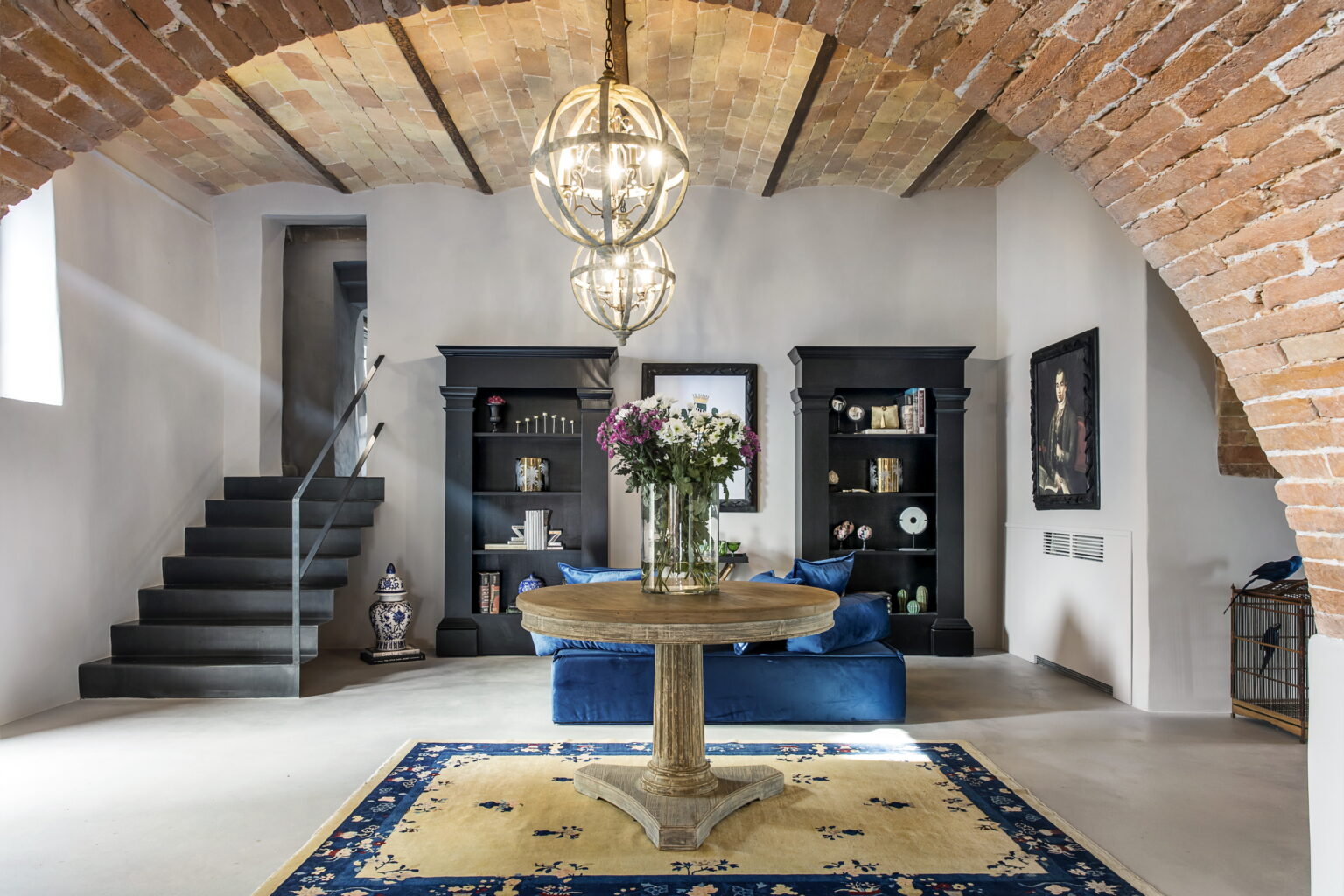Modern living room with a rustic touch, featuring a round wooden table, blue velvet couch, and black bookshelves. The space showcases exposed brick arches, stylish lighting fixtures, and decorative elements like flowers and art pieces, creating an inviting atmosphere. | Only on SopranoVillas