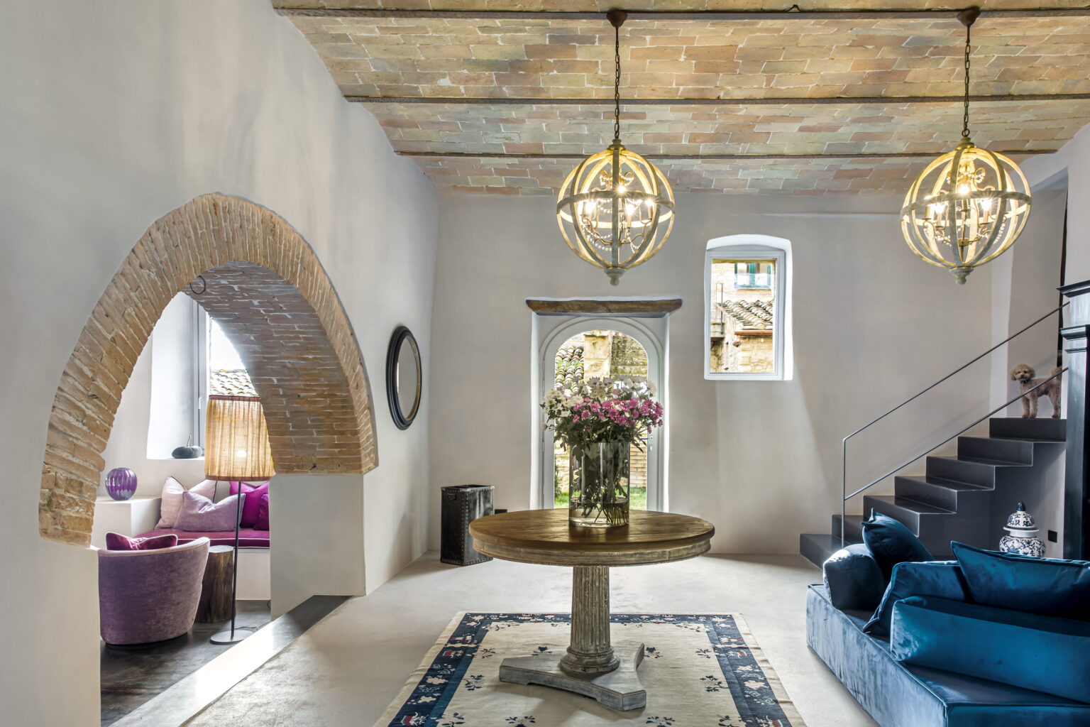 Modern interior of a stylish living space featuring a circular table with a floral arrangement, elegant pendant lights, and a staircase. The room showcases a blend of contemporary and rustic design elements, including a stone archway and a cozy seating area with purple chairs. | Only on SopranoVillas