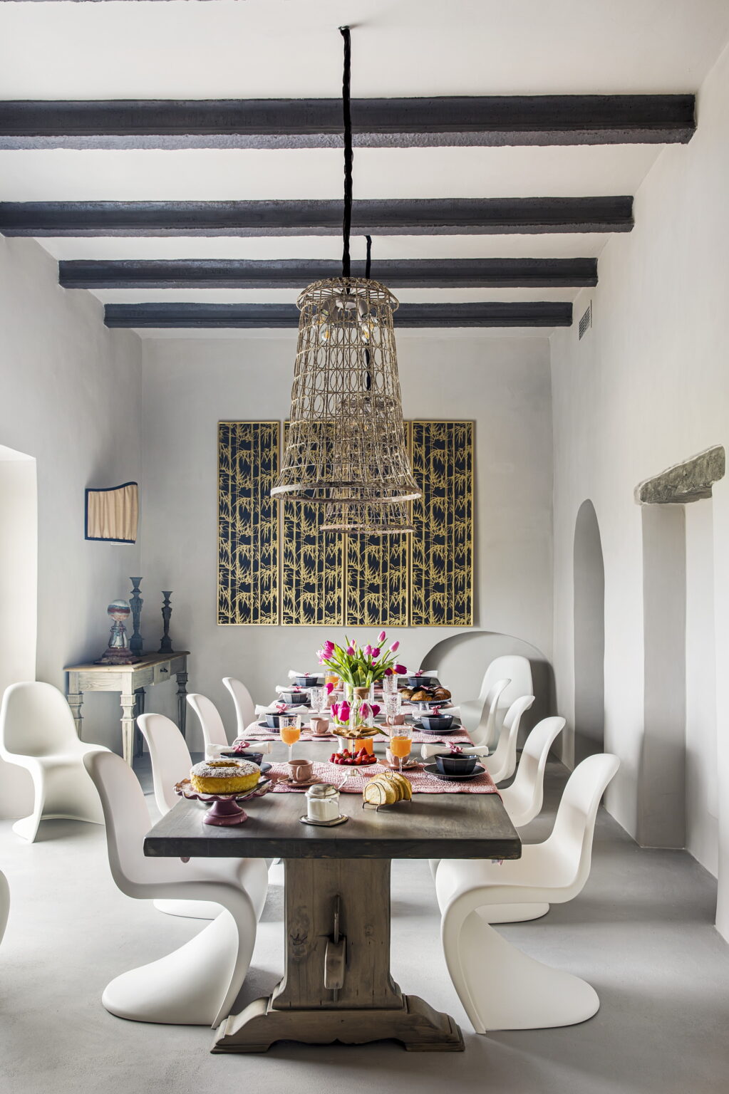 A beautifully arranged dining room featuring a large wooden table set for a meal, surrounded by modern white chairs. The table displays an array of dishes, including pastries and drinks, with a centerpiece of pink tulips. A woven pendant light hangs from the ceiling, complementing the rustic decor, while a decorative wall panel in the background adds an artistic touch. | Only on SopranoVillas