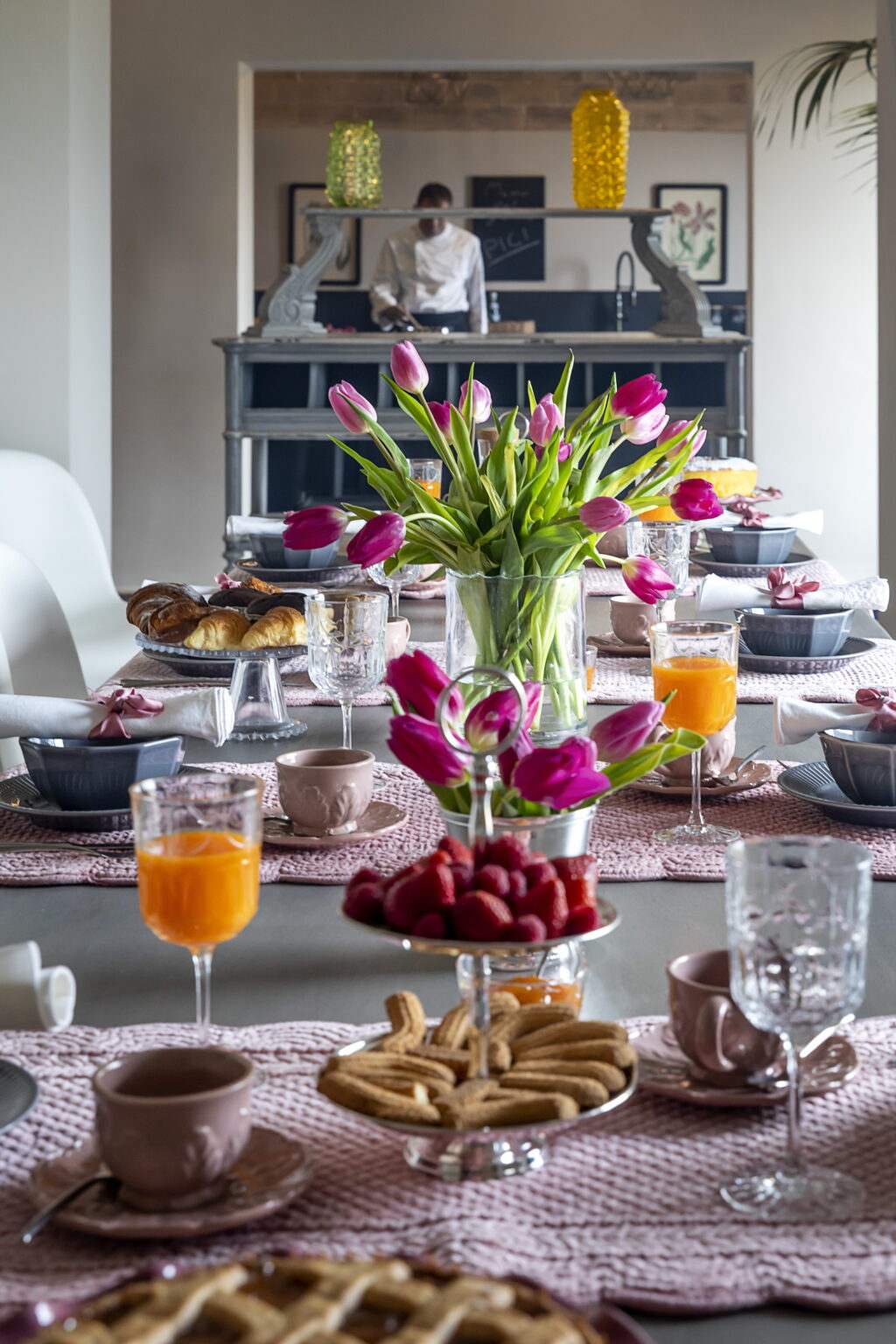 Elegant brunch setting featuring a beautifully arranged table with fresh pink tulips, assorted pastries, fruits, and beverages. A chef can be seen preparing in the background, enhancing the inviting atmosphere of the dining experience. | Only on SopranoVillas