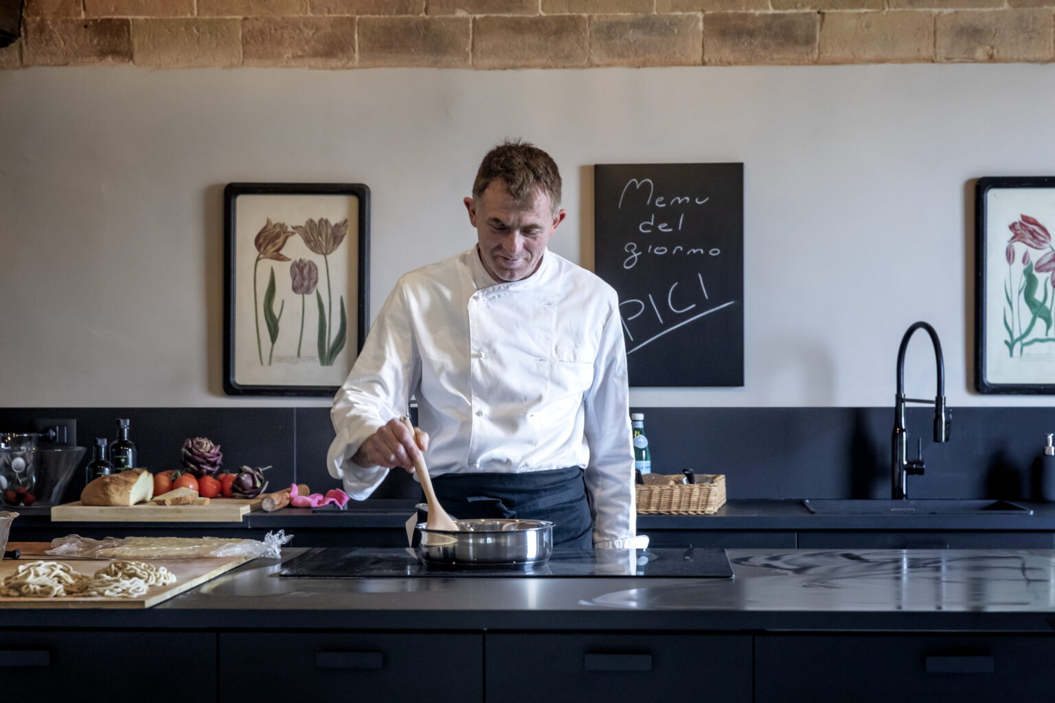 Chef cooking in a modern kitchen, stirring a pot on the stove with fresh ingredients and framed artwork in the background. | Only on SopranoVillas