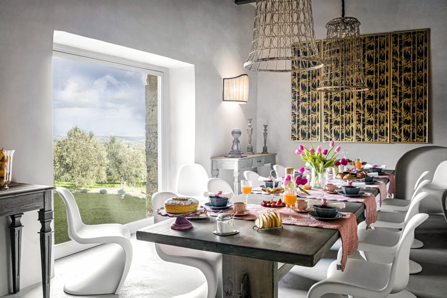 Bright and airy dining room featuring a beautifully set table with colorful dishes, flowers, and natural light streaming through a large window, overlooking a scenic landscape. | Only on SopranoVillas