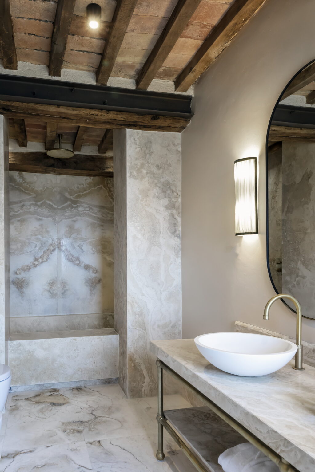 Modern bathroom interior featuring elegant marble walls, a stylish round mirror, and a minimalistic white vessel sink on a marble countertop. Exposed wooden beams add warmth to the contemporary design, creating a serene and luxurious atmosphere. | Only on SopranoVillas