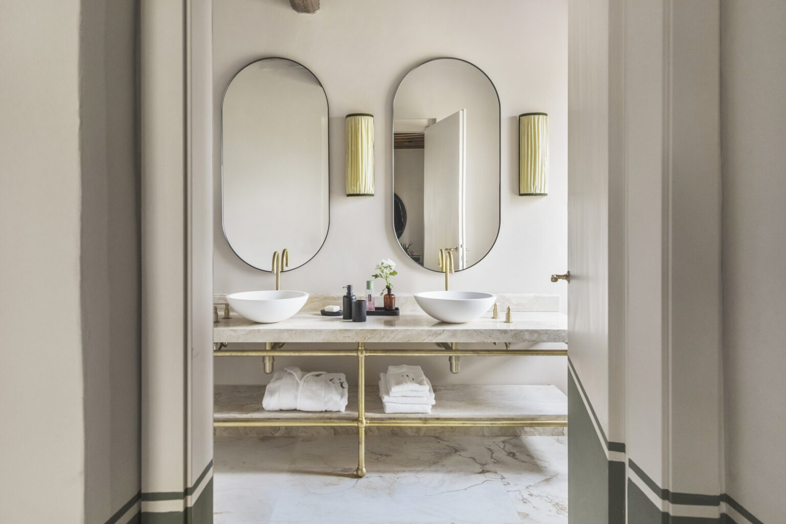 Modern bathroom interior featuring dual oval mirrors, stylish gold sconces, and a marble countertop with two elegant vessel sinks. Below, neatly arranged white towels enhance the sophisticated design. | Only on SopranoVillas