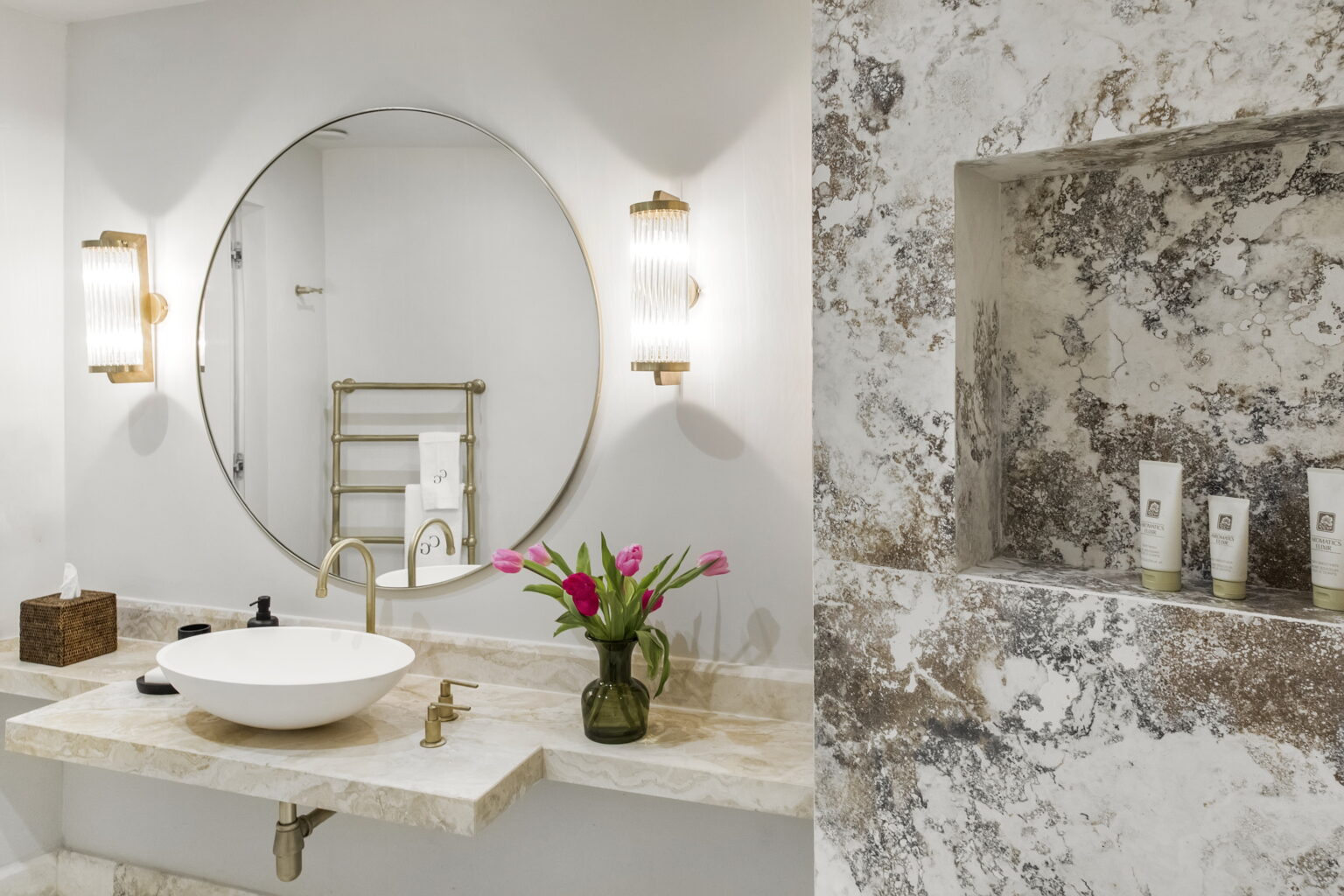 Modern bathroom featuring a round mirror, elegant wall sconces, a stylish sink on a marble countertop, and a decorative niche with skincare products and fresh flowers. | Only on SopranoVillas