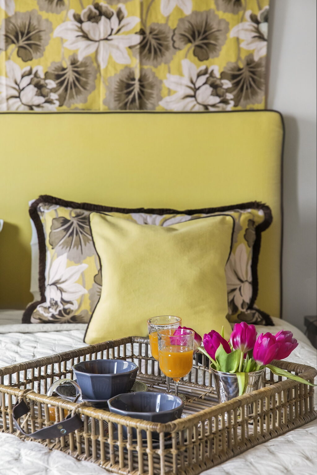 Cozy bedroom decor featuring a vibrant yellow accent pillow and a floral patterned pillow against a soft yellow wall, complemented by a glass of juice and fresh pink flowers on a bedside table. | Only on SopranoVillas
