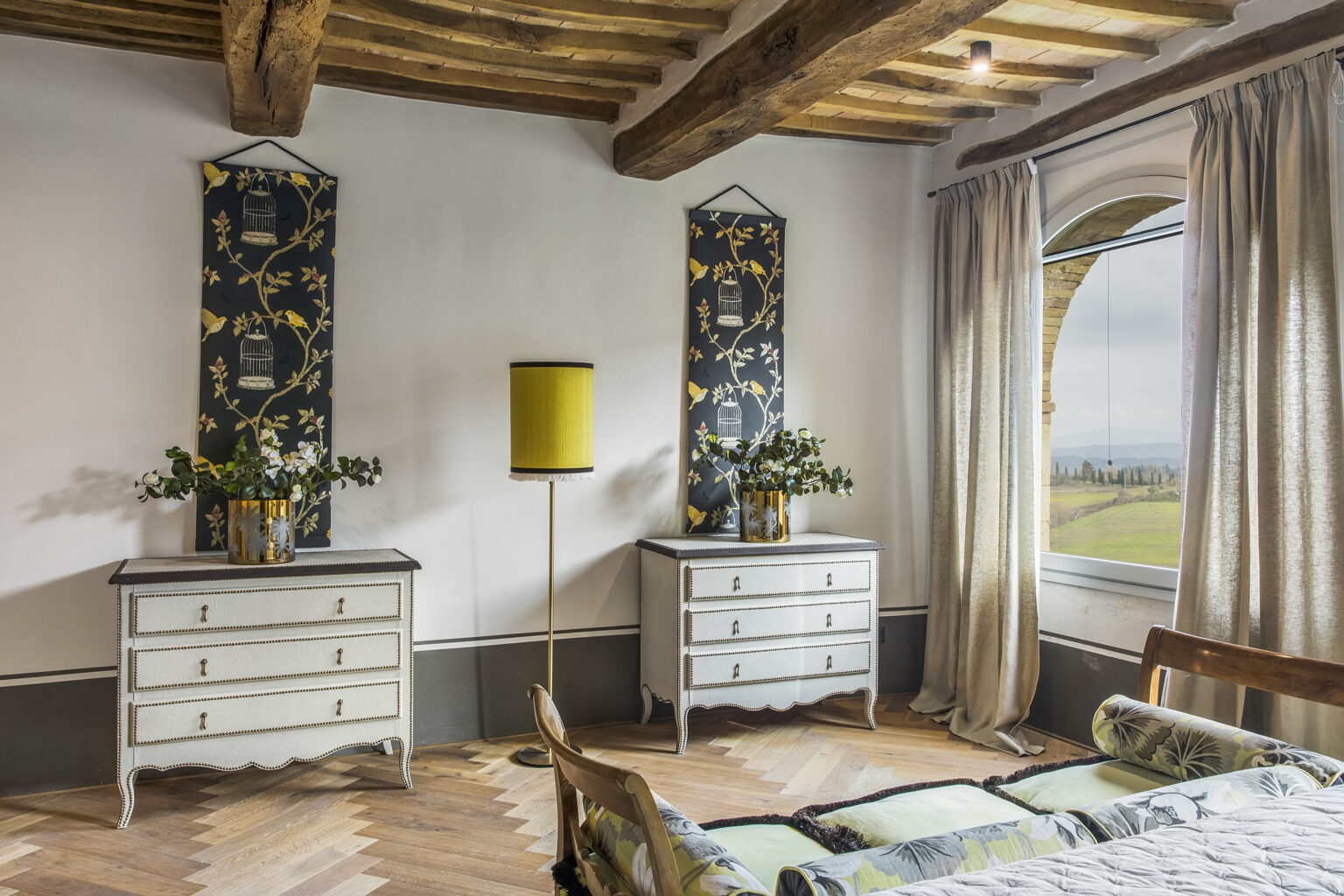 Elegant bedroom interior featuring two decorative dressers, floral wall hangings, and a modern yellow lamp. Large window offers a scenic view, complemented by natural light and wooden beams. | Only on SopranoVillas