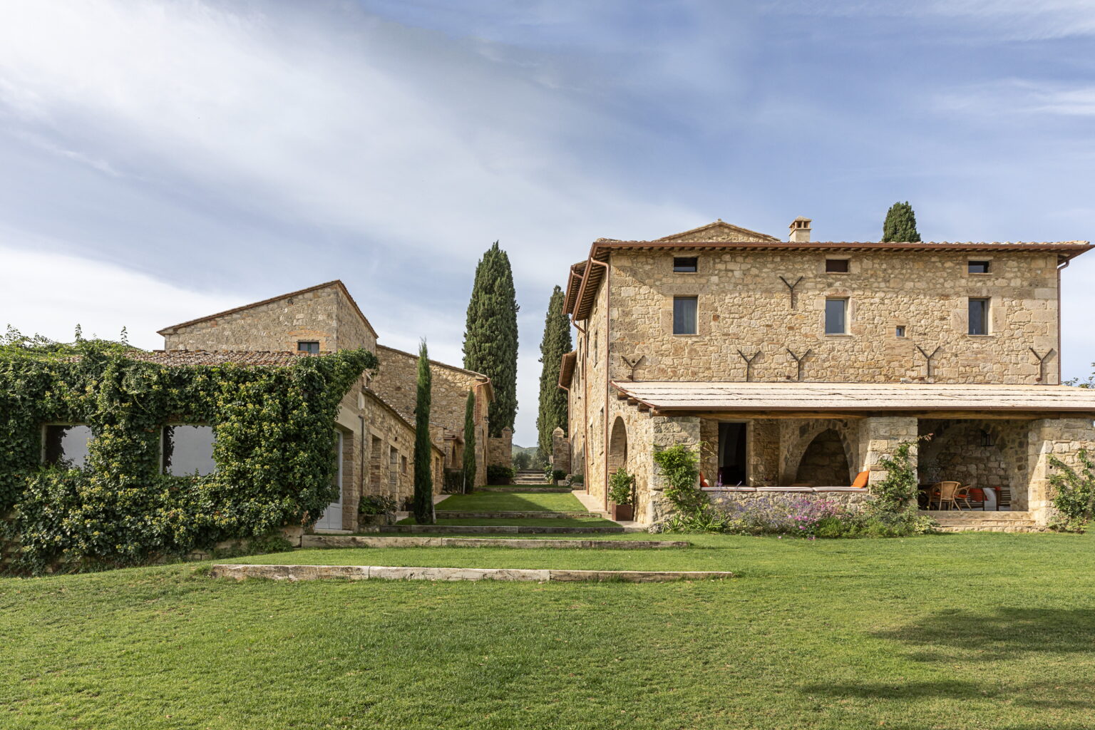 Rustic stone farmhouse surrounded by lush greenery, featuring a spacious lawn and tall cypress trees under a blue sky. Ideal for countryside retreats or vacation rentals. | Only on SopranoVillas