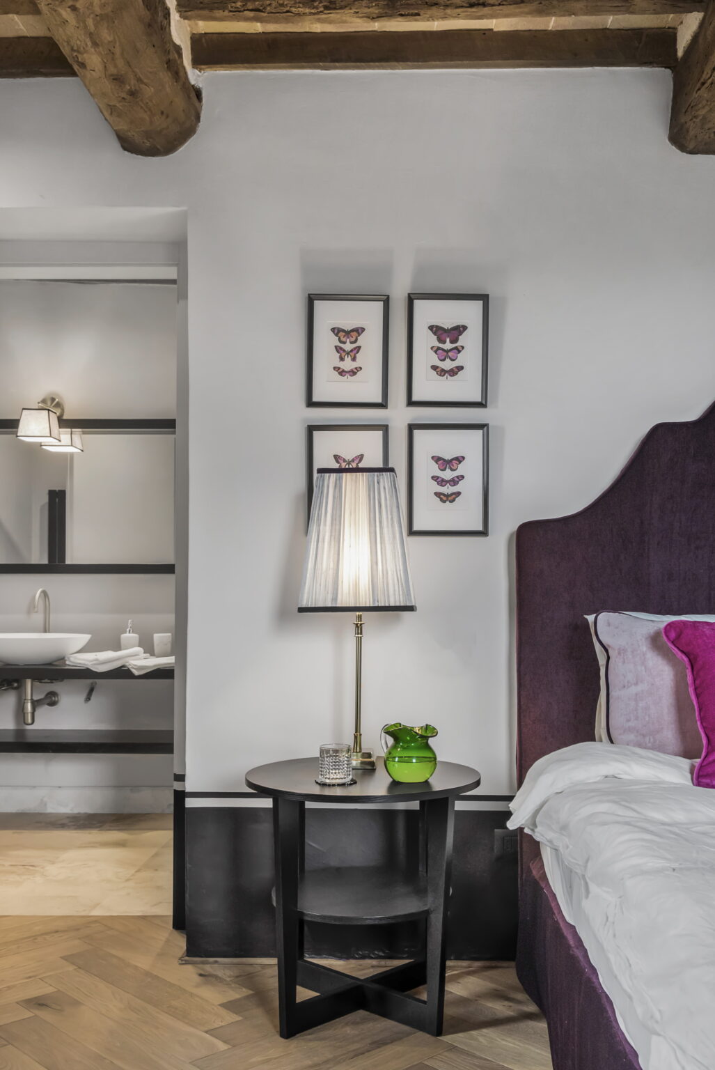 Cozy bedroom interior featuring a stylish headboard, decorative butterfly wall art, a modern lamp, and a potted plant, with an adjoining bathroom visible in the background. | Only on SopranoVillas