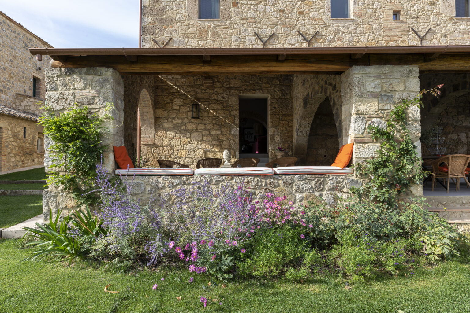 Rustic stone house with a covered porch, featuring orange cushions, surrounded by colorful flowering plants and lush green grass. | Only on SopranoVillas