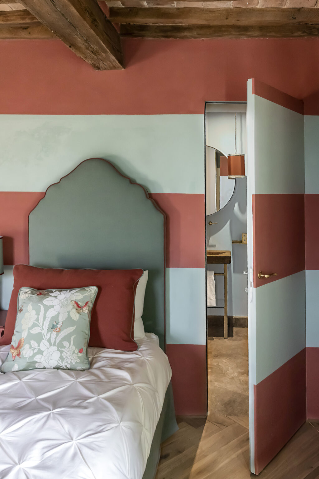 Cozy bedroom featuring a green upholstered headboard, red and teal striped walls, and a white bedspread with decorative pillows, showcasing a doorway leading to a stylish bathroom with a round mirror and modern lighting. | Only on SopranoVillas