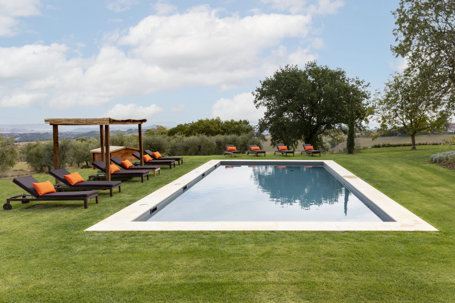 Modern outdoor swimming pool surrounded by lush green grass, featuring comfortable lounge chairs with orange cushions and wooden pergolas, set against a scenic landscape under a partly cloudy sky. | Only on SopranoVillas