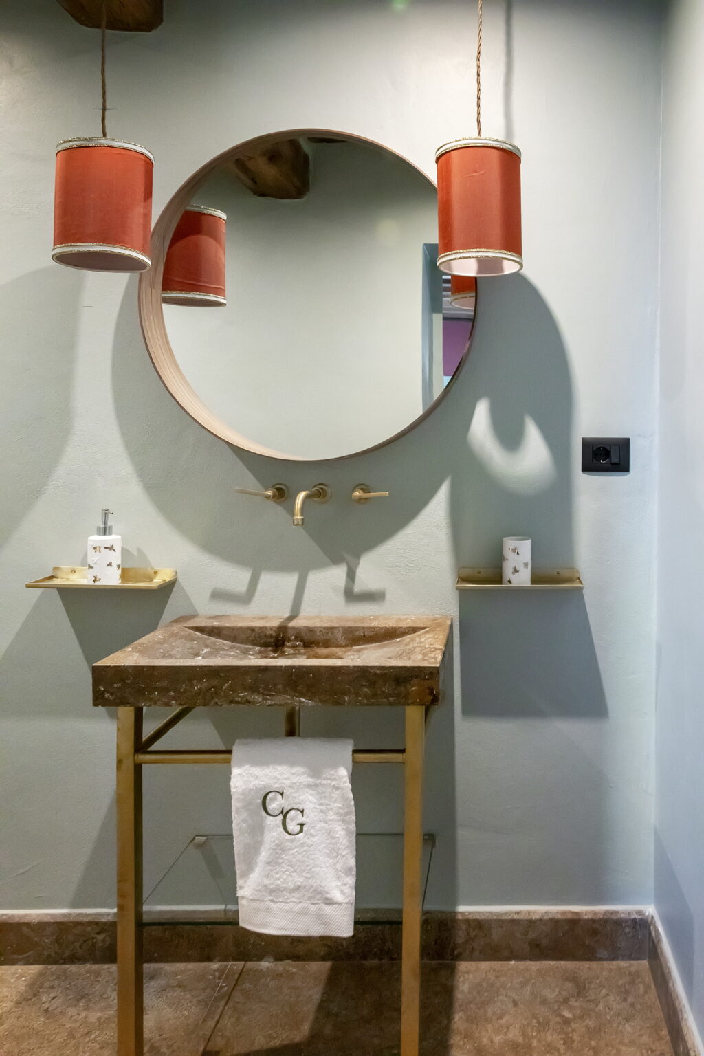 Modern bathroom design featuring a stylish round mirror above a unique stone sink, complemented by gold faucets and minimalist wall shelves with decorative items. | Only on SopranoVillas