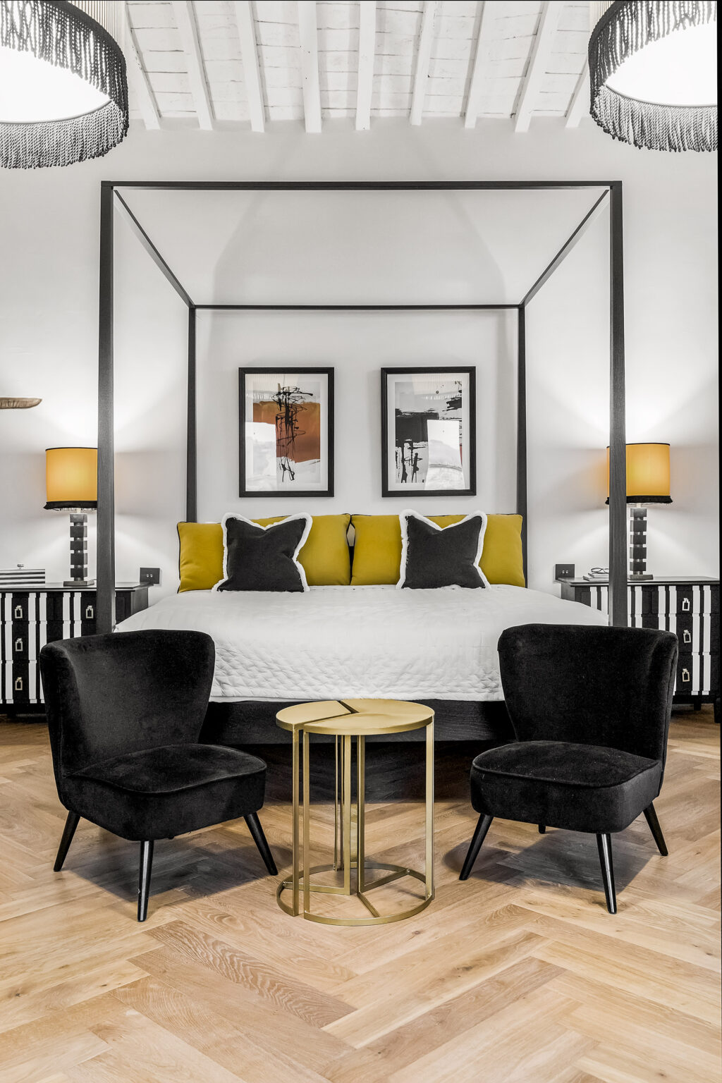 Modern bedroom featuring a four-poster bed with yellow and black accent pillows, framed artwork on the walls, stylish black chairs, and contemporary bedside lamps, creating a cozy and elegant atmosphere. | Only on SopranoVillas