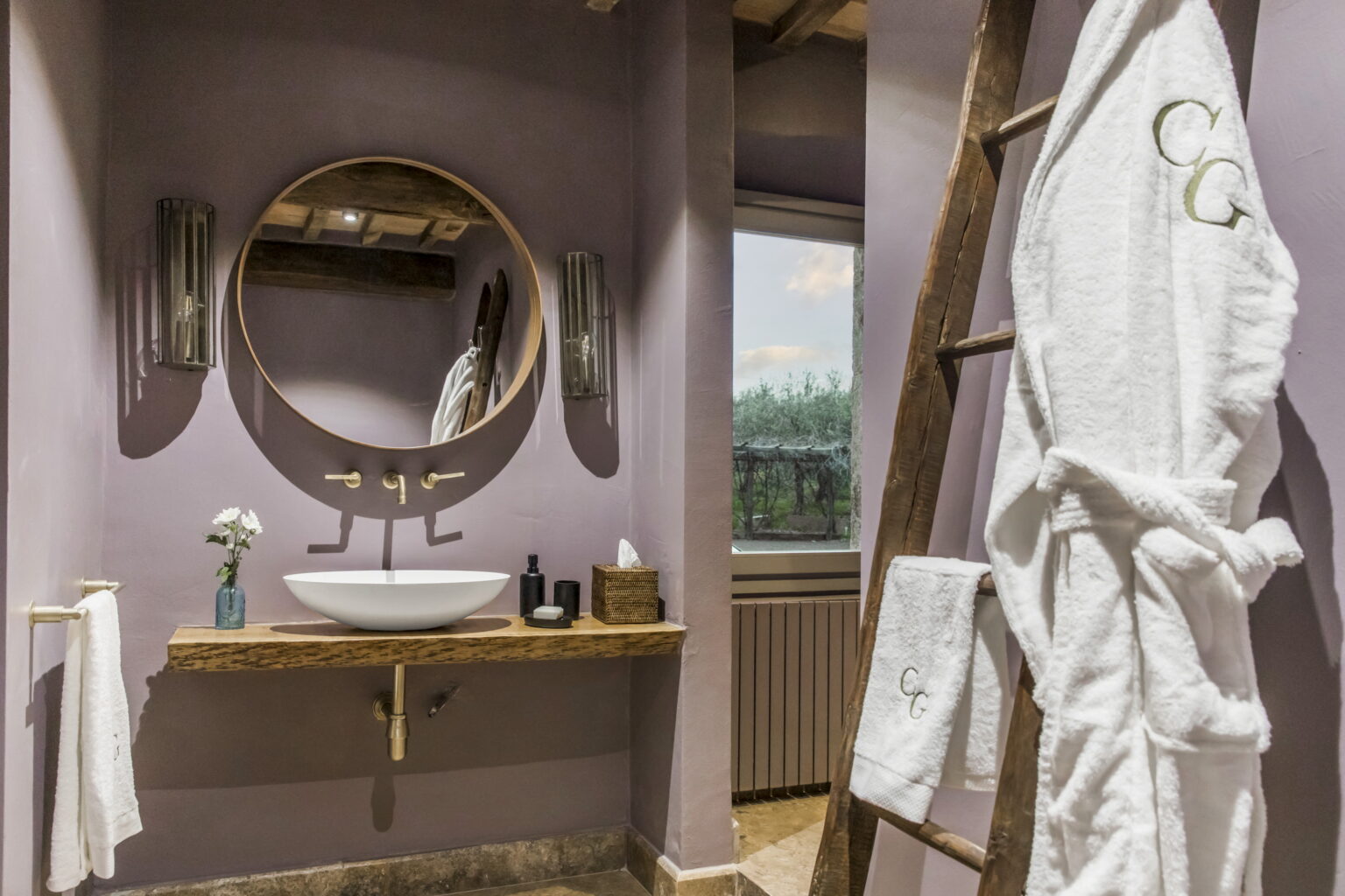 Modern bathroom interior featuring a round mirror, a stylish sink on a wooden countertop, and plush white bathrobes hanging on a ladder. Soft purple walls and natural light enhance the relaxing atmosphere. | Only on SopranoVillas