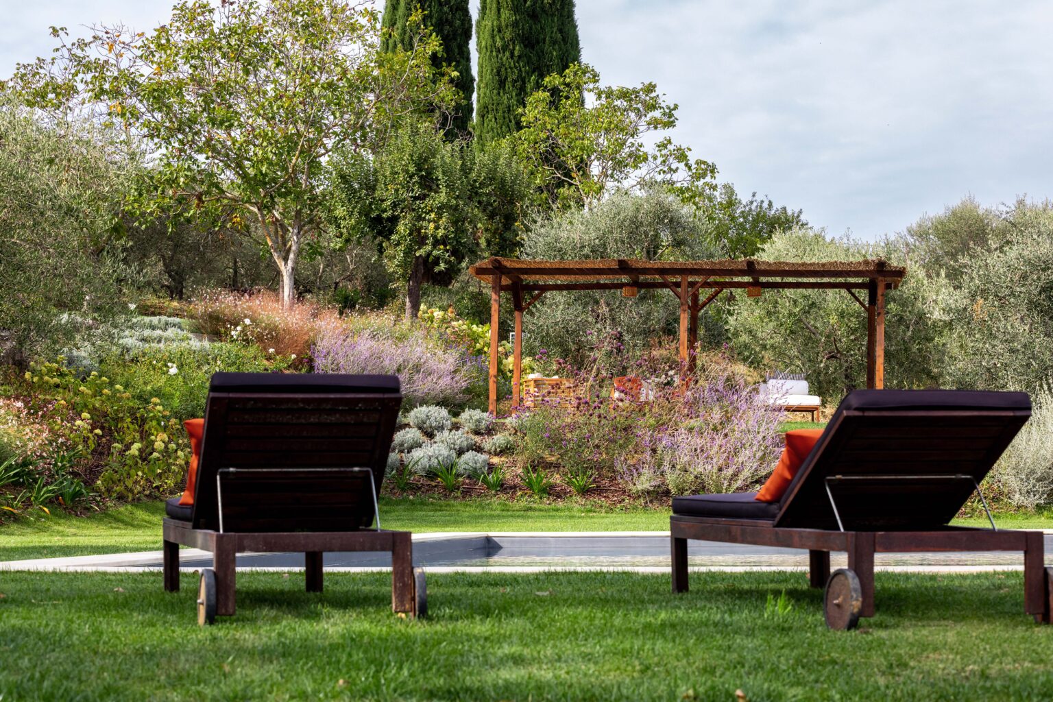Two wooden lounge chairs with orange cushions facing a serene garden featuring lush greenery, flowering plants, and a wooden pergola, creating a tranquil outdoor relaxation space. | Only on SopranoVillas