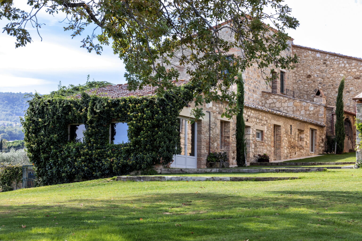 Rustic stone villa surrounded by lush greenery and rolling hills in a picturesque landscape. The building features ivy-covered walls and large windows, offering a serene countryside retreat. | Only on SopranoVillas
