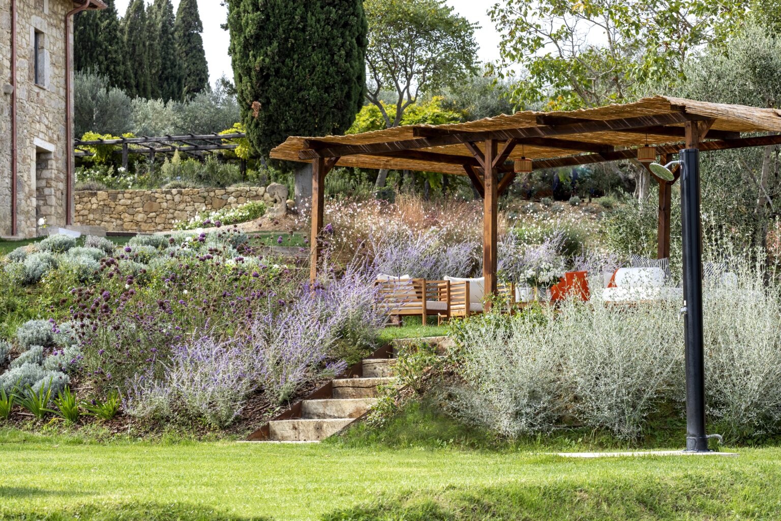 Wooden pergola surrounded by lush lavender and greenery in a tranquil garden setting, featuring outdoor seating and stone steps leading to the landscape. | Only on SopranoVillas