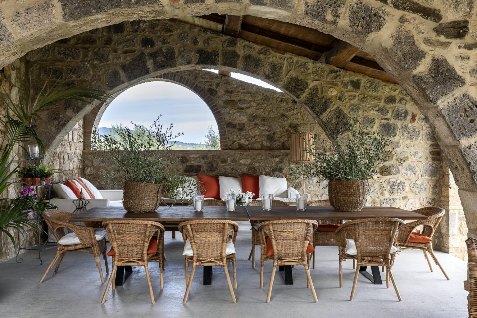 Rustic dining area featuring a long wooden table surrounded by wicker chairs, adorned with potted plants and decorative cushions, set within a stone archway that frames a scenic view. | Only on SopranoVillas
