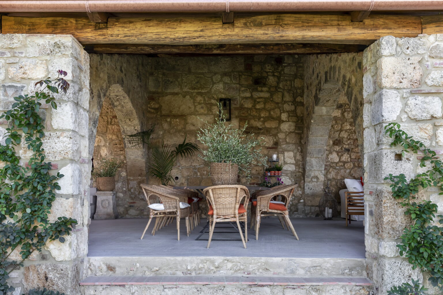 Cozy outdoor dining area with rustic stone walls, featuring a wooden table and wicker chairs, surrounded by greenery and natural light. Ideal for al fresco gatherings. | Only on SopranoVillas