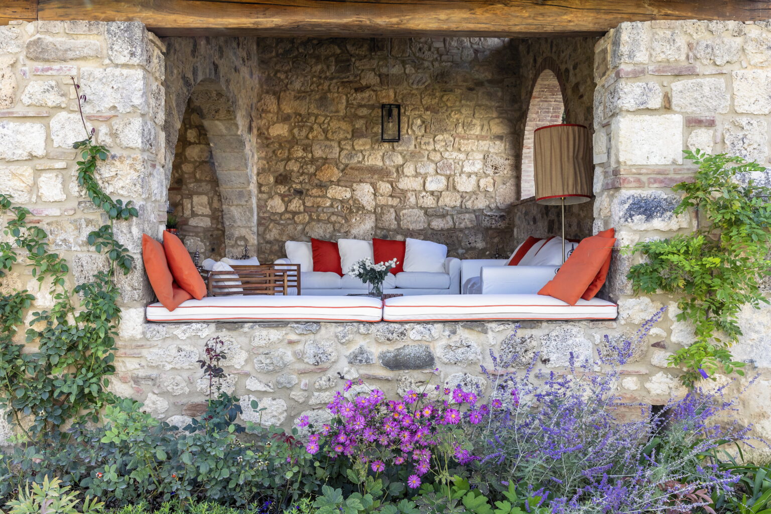 Cozy outdoor seating area featuring white sofas with orange cushions, set against a rustic stone wall. Decorative plants and colorful flowers enhance the inviting atmosphere. Perfect for relaxation and enjoying nature. | Only on SopranoVillas