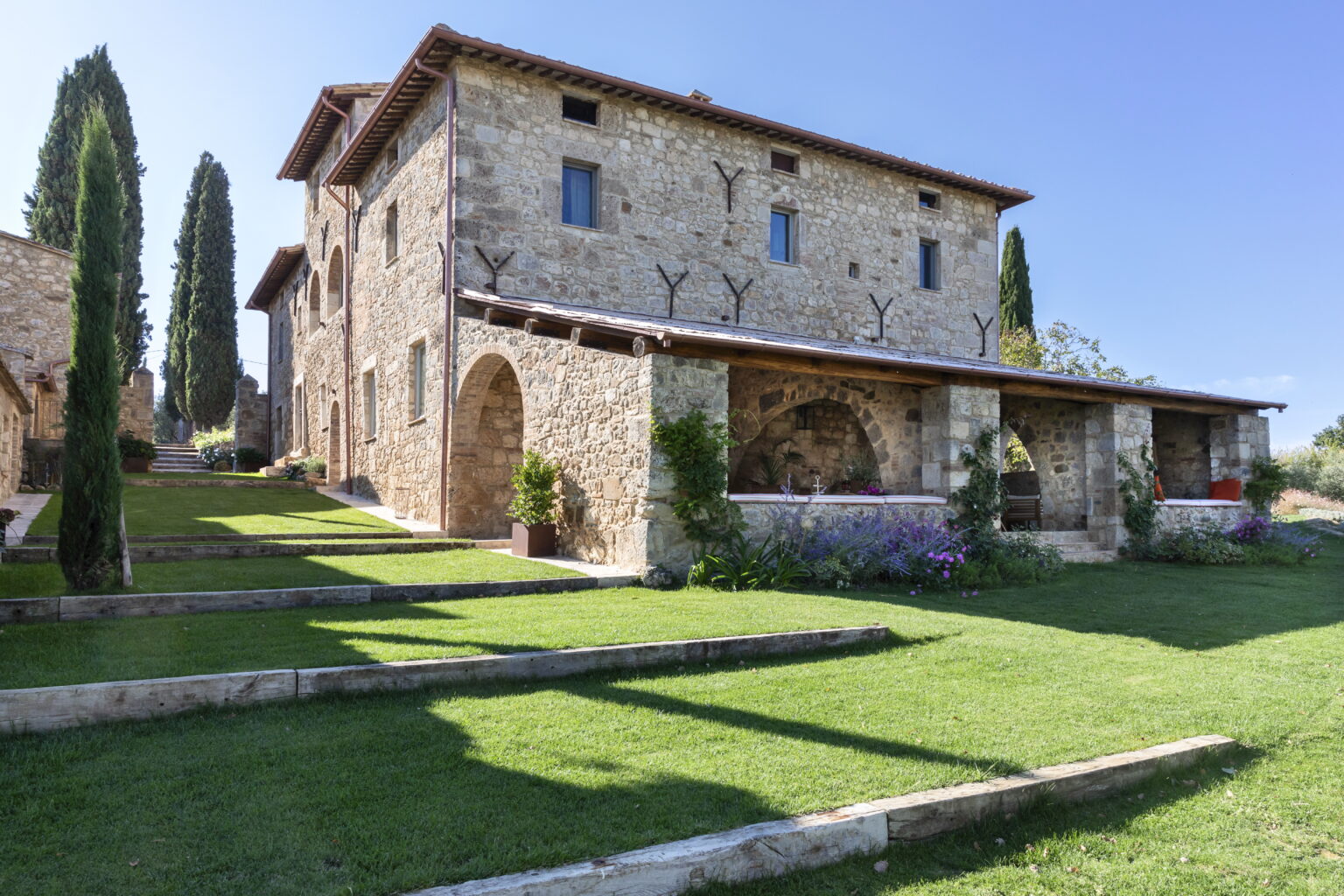 Stone villa with a rustic design, surrounded by lush green lawns and cypress trees under a clear blue sky. Ideal for luxury vacation rentals in a picturesque countryside setting. | Only on SopranoVillas