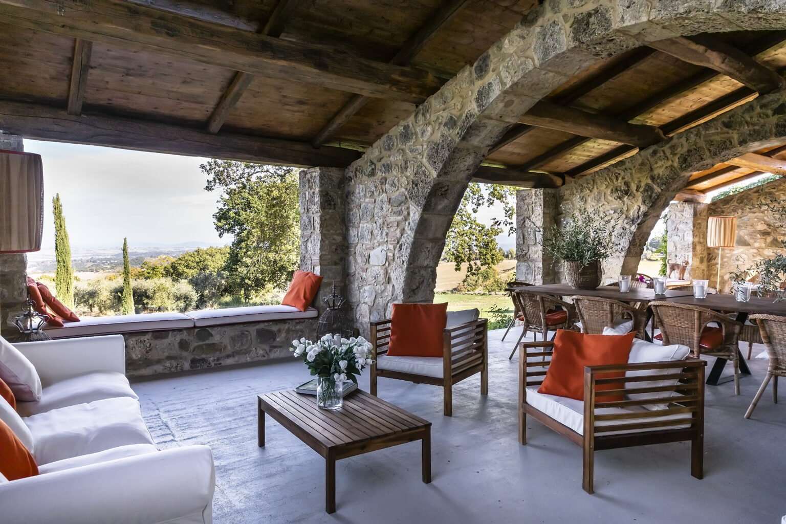 Cozy outdoor living space featuring a stone patio with wooden beams, comfortable seating arrangements, and a dining area, all surrounded by scenic views of lush greenery and rolling hills. | Only on SopranoVillas