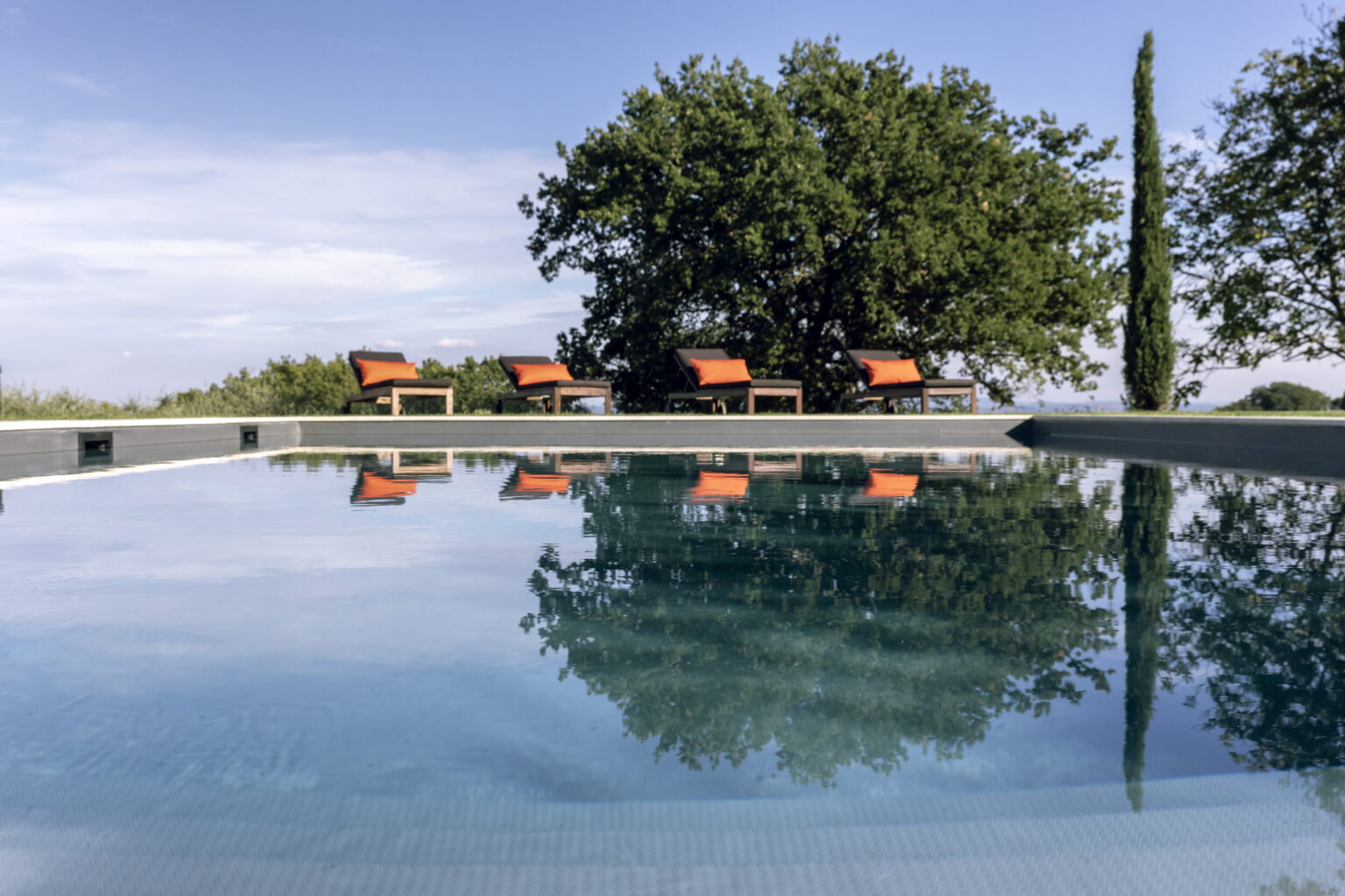 Tranquil swimming pool reflecting blue sky and surrounding greenery, with stylish loungers featuring orange cushions in the background, ideal for relaxation and outdoor leisure. | Only on SopranoVillas