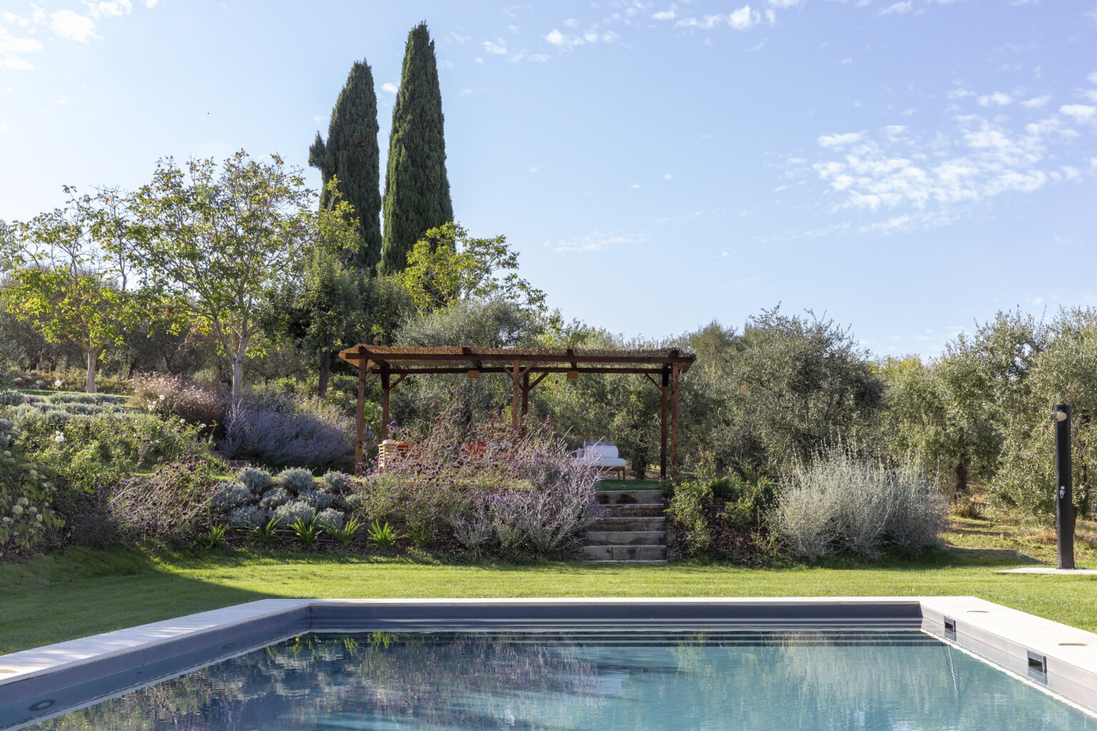 Serene outdoor pool area with a pergola surrounded by lush greenery and flowering plants, set against a clear blue sky and tall cypress trees in the background. | Only on SopranoVillas