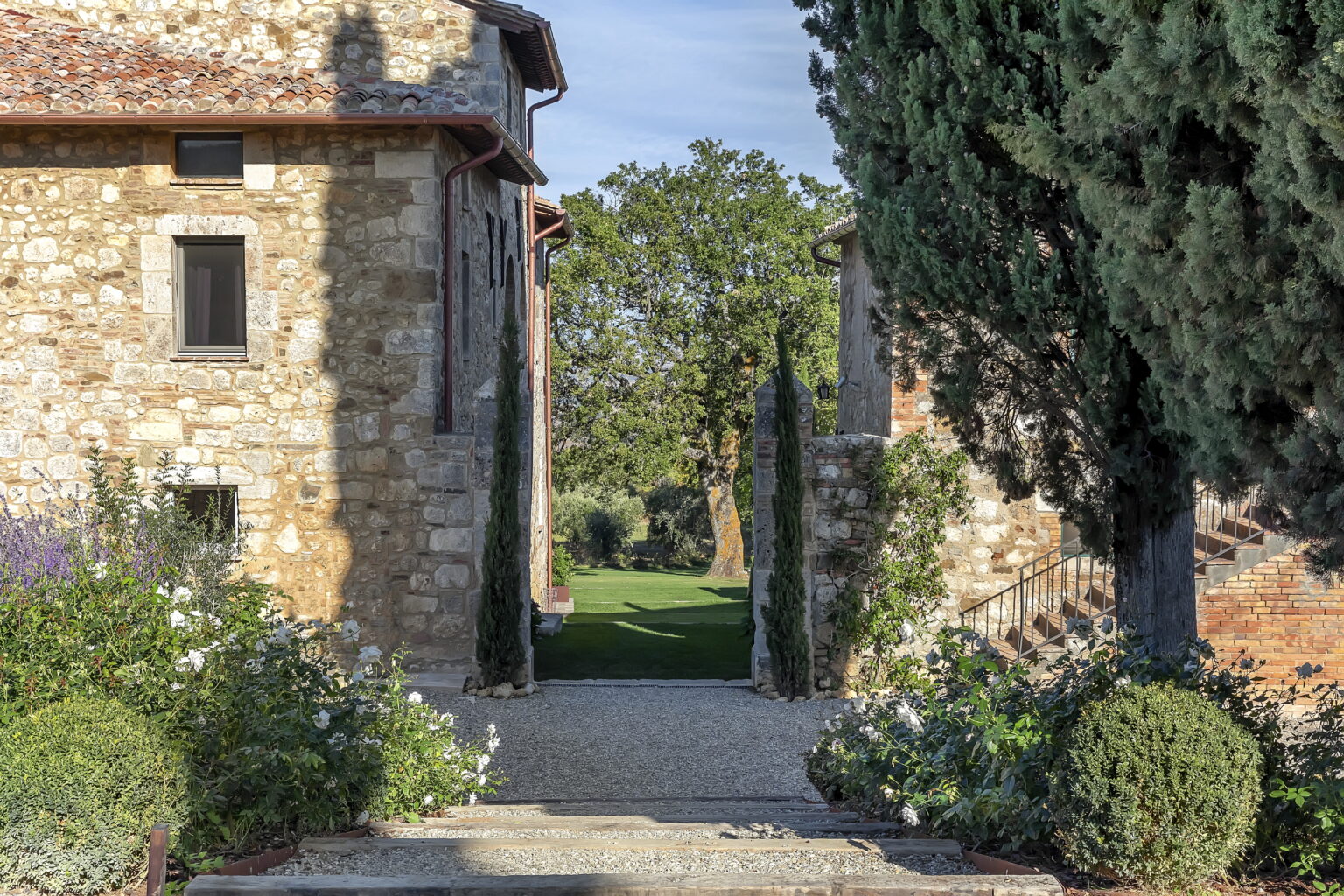 Scenic pathway between rustic stone buildings, lined with green shrubs and cypress trees, leading to a lush garden in a tranquil countryside setting. | Only on SopranoVillas