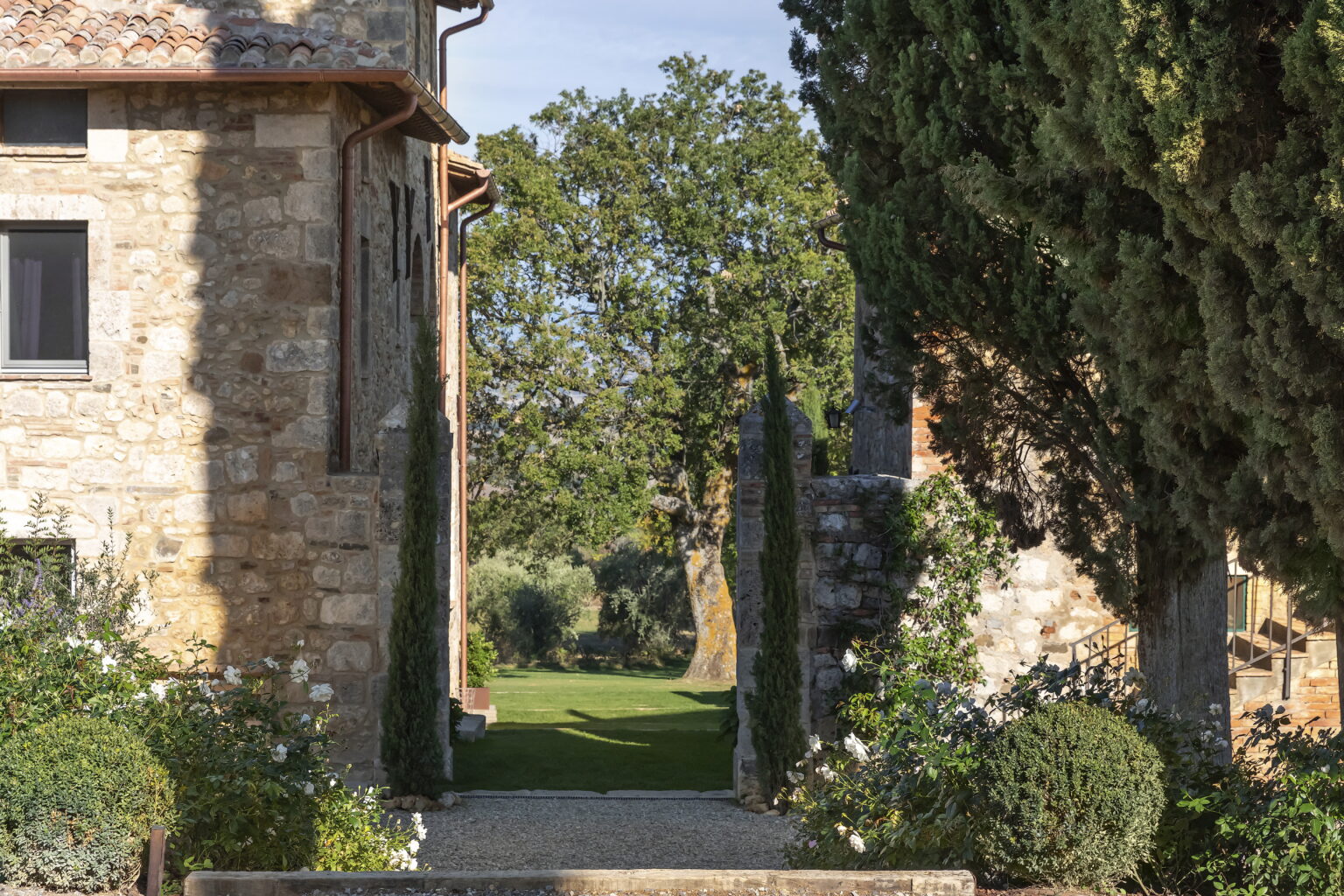 Stone pathway between two rustic buildings, leading to a lush green garden with trees and flowering shrubs under a clear blue sky. | Only on SopranoVillas