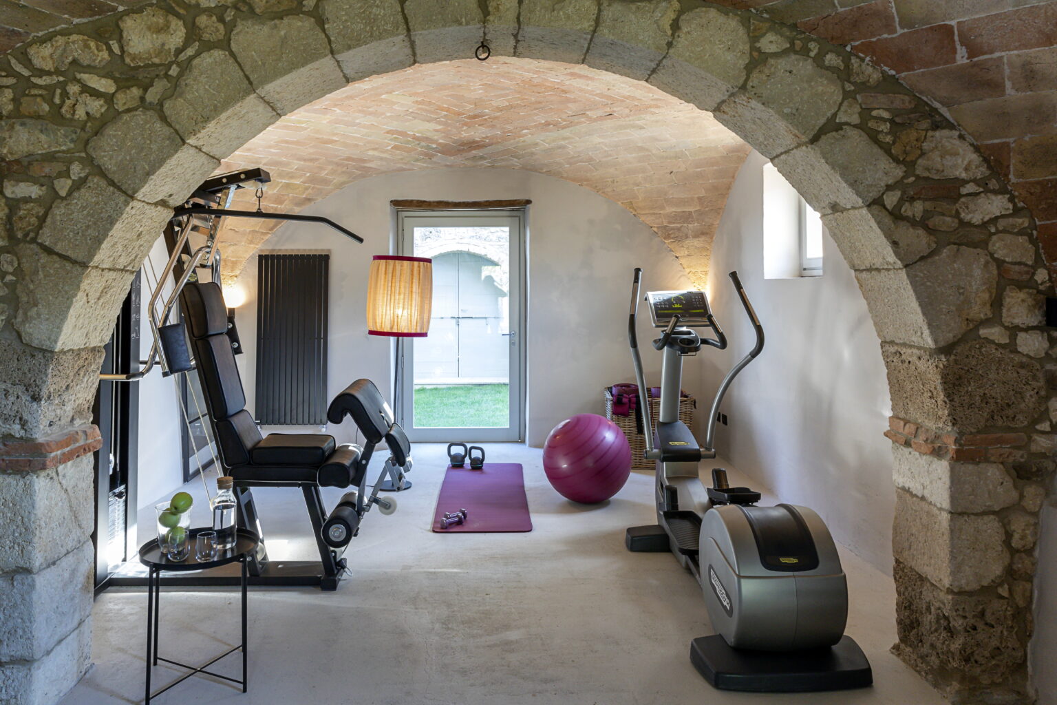 Home gym featuring modern fitness equipment, including a multi-station weight machine, elliptical trainer, and exercise ball, set in a stylish room with an arched stone ceiling and natural light. | Only on SopranoVillas