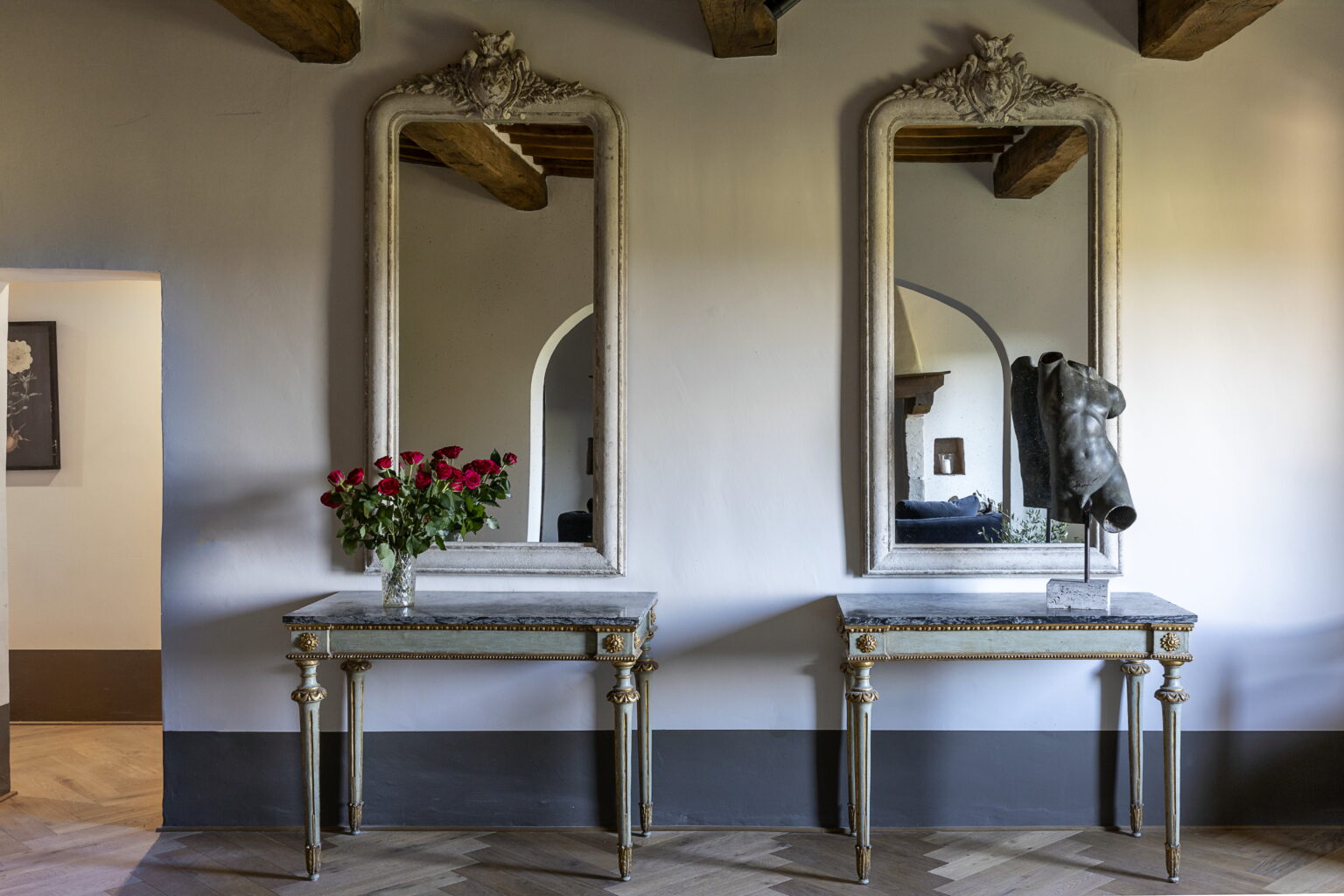 Elegant interior featuring two ornate mirrors above decorative tables, adorned with a bouquet of red roses and a sculptural torso piece. The room showcases classic design elements with wooden beams and stylish decor, creating a sophisticated ambiance. | Only on SopranoVillas