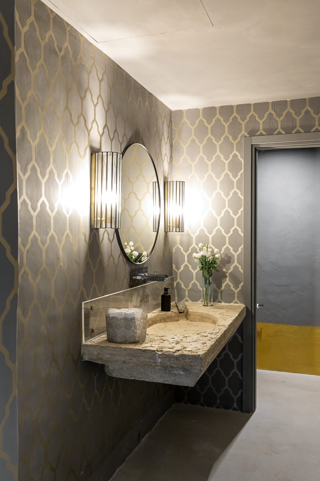 Modern bathroom interior featuring a stylish vanity with a circular mirror, elegant wall sconces, and a decorative backsplash. The space is adorned with a vase of fresh flowers and neutral color tones, creating a sophisticated atmosphere. | Only on SopranoVillas