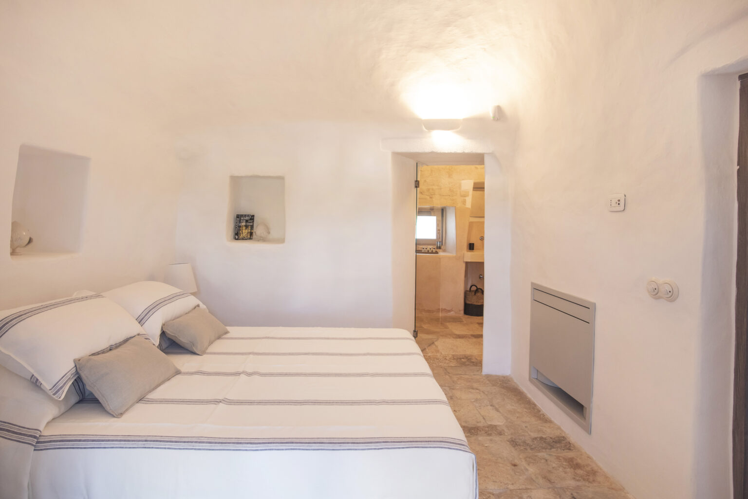 Modern and minimalist bedroom featuring a large bed with neutral-colored linens, soft lighting, and a view into a connected bathroom. The white walls and stone flooring create a clean and airy atmosphere, perfect for relaxation. | Only on SopranoVillas