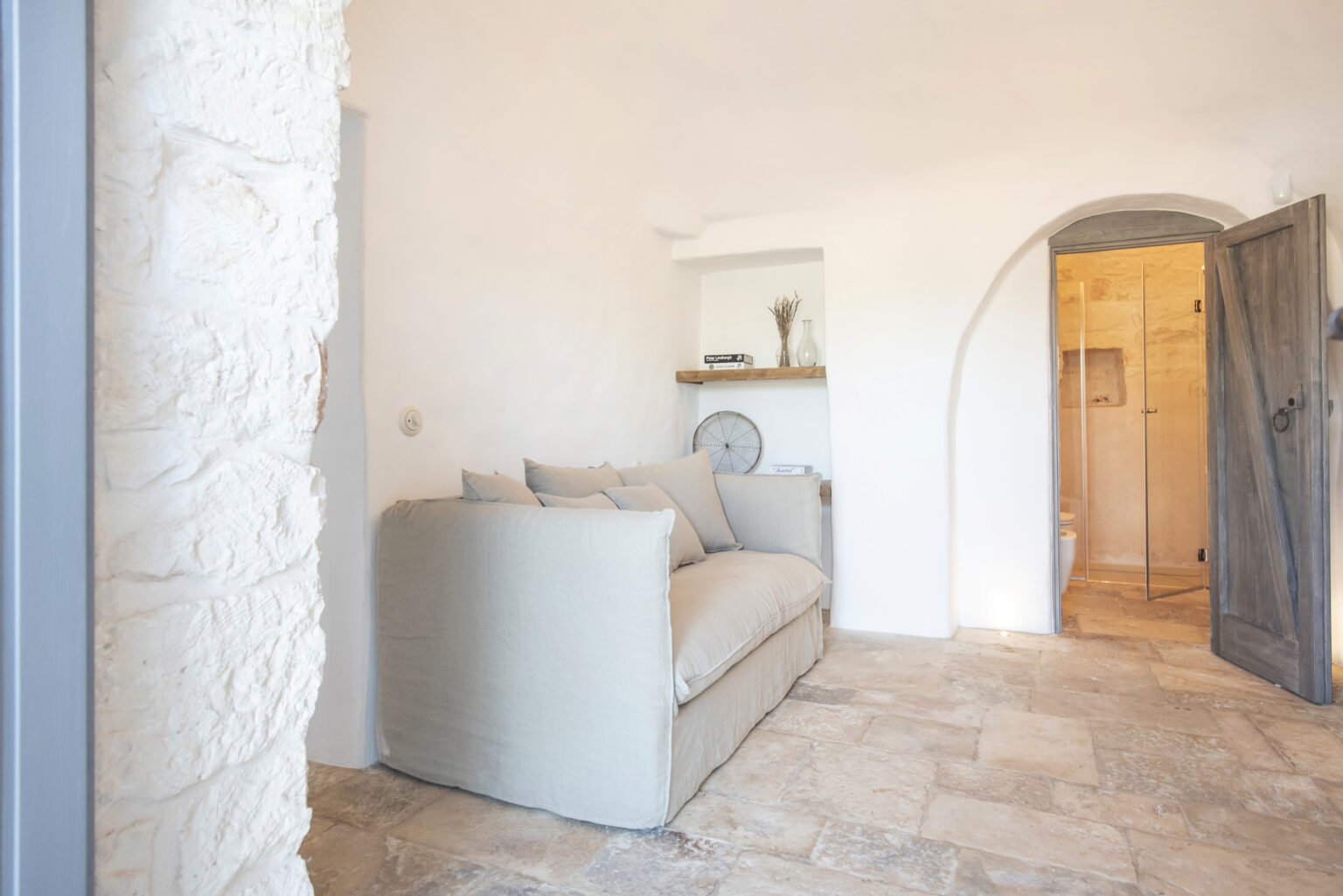 Cozy living room with a light-colored sofa, stone flooring, and an arched doorway leading to a bathroom. The space features minimalist decor and natural light. | Only on SopranoVillas