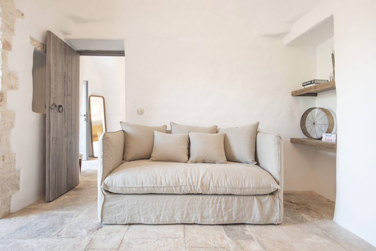 Cozy living room featuring a beige sofa with multiple cushions, set against a minimalist white wall. Natural light floods the space, highlighting wooden accents and a stylish shelf with decorative items. | Only on SopranoVillas