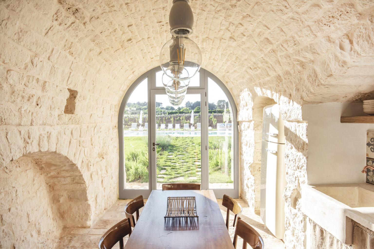 Interior view of a rustic dining area featuring a wooden table, stone walls, and a large arched window that offers a scenic view of the outdoors, highlighting natural light and a cozy atmosphere. | Only on SopranoVillas