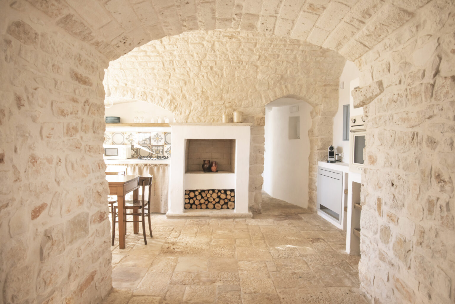 Interior view of a rustic kitchen featuring stone archways, a wooden dining table with chairs, a fireplace with stacked logs, and a well-equipped kitchen area with decorative tiles and modern appliances. | Only on SopranoVillas