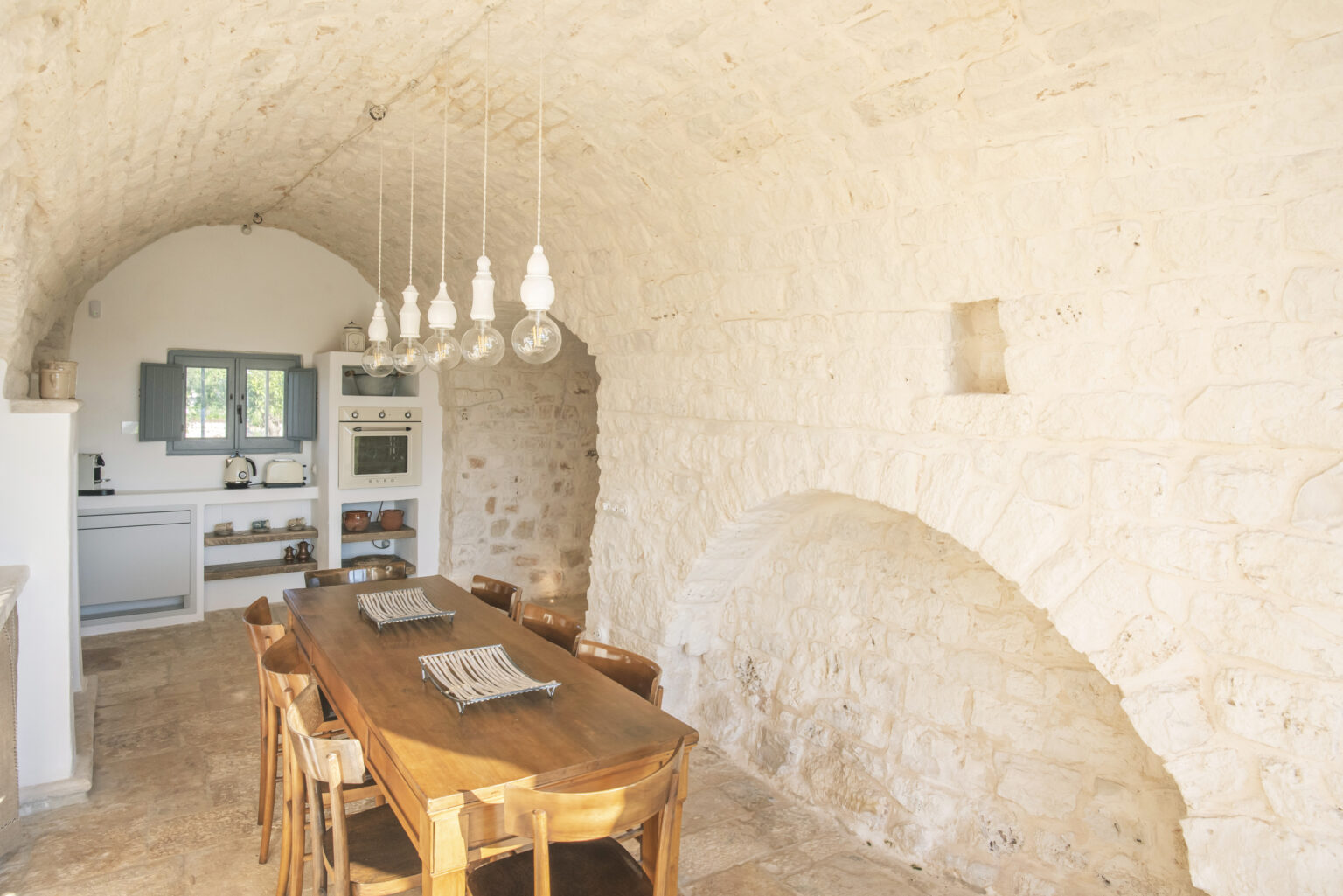 Rustic kitchen interior featuring an arched stone ceiling, wooden dining table, and modern light fixtures, with white cabinetry and a window allowing natural light. | Only on SopranoVillas